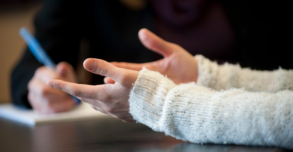 Two people at a meeting; one gesturing with hands, the other taking notes